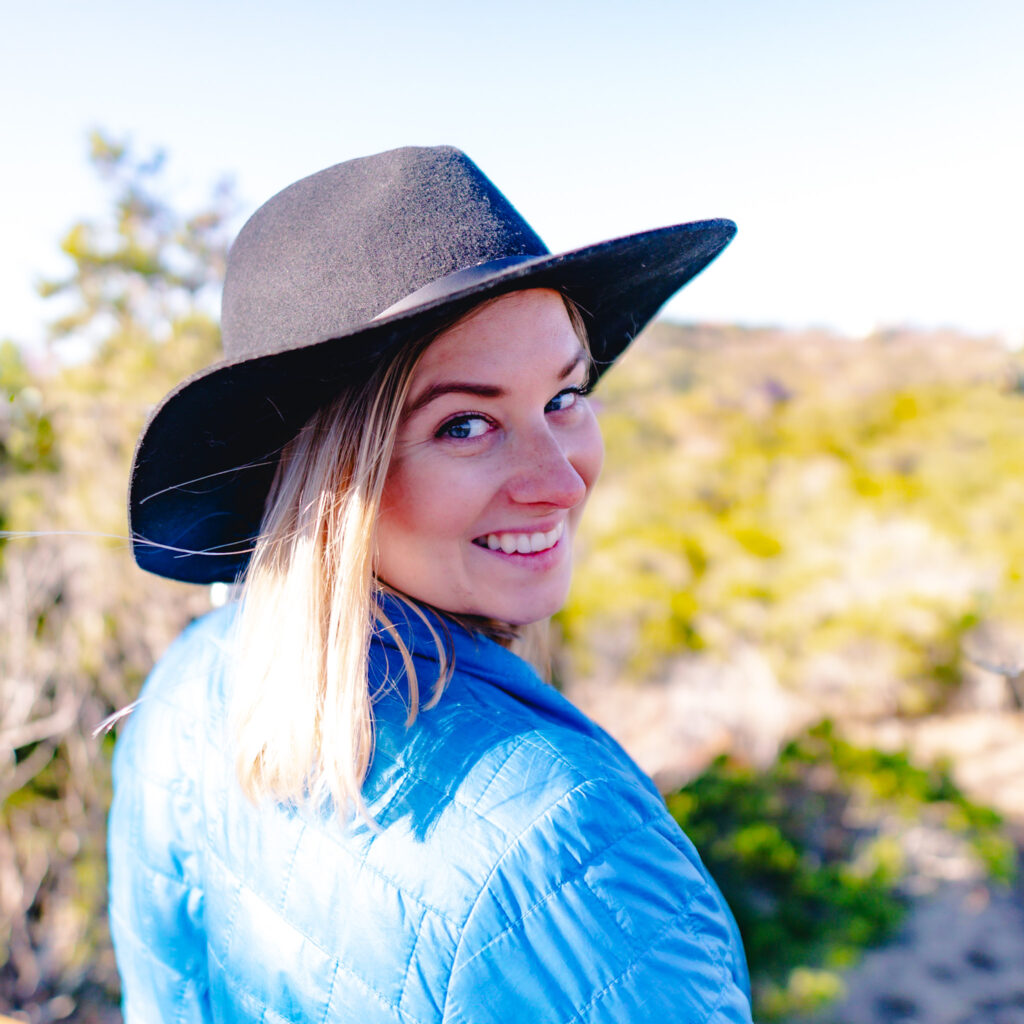 A photo of Blair, a white woman with shoulder-length blonde hair and blue eyes. She is wearing a black felt hat and blue nano puff jacket and smiles at the viewer, looking over her right shoulder. Rolling hills with spots of greenery can be discerned in the background.